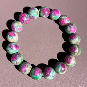 May include: A beaded bracelet featuring round gemstones. The stones are a mix of colors, with a base of light green and cream, and vibrant pink and red accents. The bracelet is strung together, forming a circular shape.