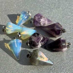 May include: A collection of crystal pendulums in various colors. The pendulums are teardrop-shaped with silver-colored metal loops. Colors include opalescent white, blue, purple, and iridescent gray. The crystals are arranged on a textured, light gray surface.