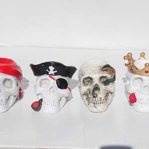 May include: Four white skull figurines with different decorative details. One skull wears a red bandana, another a black pirate hat with an eye patch, another has a green snake wrapped around it, and the last one wears a gold crown with red jewels.
