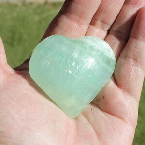 May include: A heart-shaped, pale green stone. The polished surface of the stone has a translucent quality, with subtle variations in color. The heart is held in an open hand, with a green grassy background.