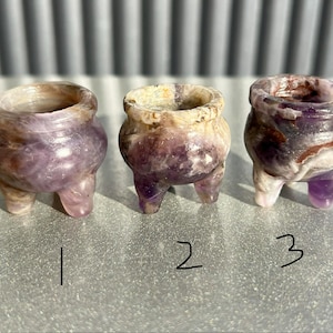May include: Three small, amethyst-colored cauldrons with three legs each. The cauldrons have a rounded body and a wide opening. The amethyst color varies from light to dark purple with some white and brown marbling. Numbers 1, 2, and 3 are written below each cauldron.