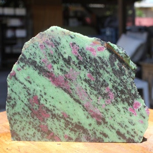 May include: A cut and polished gemstone with a vibrant green base color, interspersed with black and deep red mineral inclusions. The stone's surface is textured, revealing its natural composition. The cut edge shows the layered structure of the stone.
