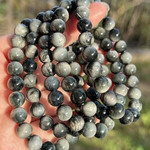 May include: A collection of multiple bracelets made of round, polished beads. The beads are primarily shades of gray and black, with a marbled appearance. The bracelets are held in a hand, with the background out of focus.