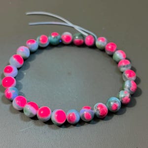 May include: A beaded bracelet featuring round beads in shades of pink, blue, and green. The beads have a marbled appearance with a bright pink accent. The bracelet is strung on a clear elastic cord.