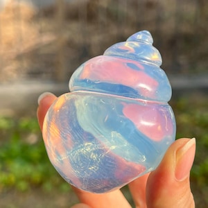 May include: A translucent, opalescent crystal sculpture in the shape of a seashell. The crystal has swirls of pink, blue, and white, with a polished, smooth surface. The sculpture is held in a hand, with a blurred green background.