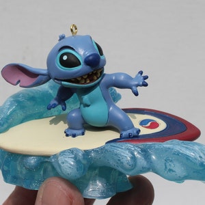May include: Blue Stitch character figurine riding a surfboard on a blue wave. The surfboard has red, white, and blue accents.