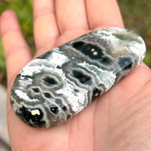 May include: An oval-shaped polished stone featuring a complex pattern of deep forest green, charcoal black, and bright white swirls. The surface displays distinct dark circular inclusions with small yellow centers, creating a natural eye-like effect.