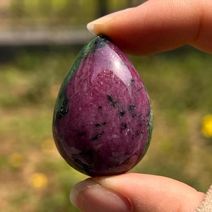 May include: A polished, teardrop-shaped gemstone featuring a deep magenta and purple base color with scattered black mineral inclusions and streaks of dark green along the edges, showcasing a smooth, reflective surface and natural stone texture.