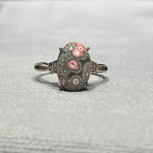 Peut inclure: Une bague argent&eacute;e orn&eacute;e d'une pierre cabochon ovale &agrave; base grise avec des motifs circulaires roses et blancs distincts. L'anneau en m&eacute;tal comporte de petites pierres transparentes incrust&eacute;es sur les c&ocirc;t&eacute;s de la monture centrale.