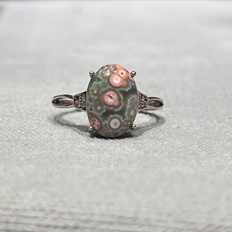 Peut inclure: Une bague argent&eacute;e orn&eacute;e d'une pierre cabochon ovale &agrave; base grise avec des motifs circulaires roses et blancs distincts. L'anneau en m&eacute;tal comporte de petites pierres transparentes incrust&eacute;es sur les c&ocirc;t&eacute;s de la monture centrale.