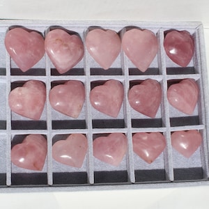 May include: A collection of 16 pink heart-shaped rose quartz stones arranged in a grid pattern in a white box.