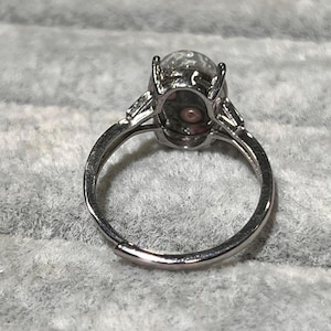 Peut inclure: Une bague argent&eacute;e orn&eacute;e d'une pierre ovale translucide avec des inclusions sombres et un motif circulaire central. L'anneau en m&eacute;tal pr&eacute;sente un sertissage &agrave; griffes pour la pierre et une ouverture &agrave; la base, indiquant un design ajustable.