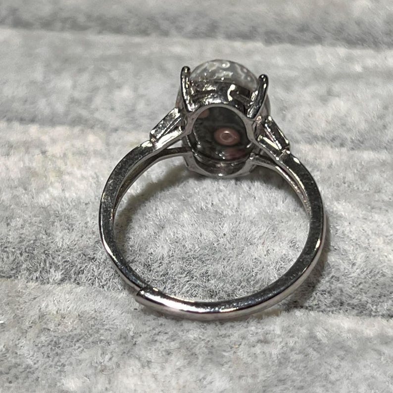 Peut inclure: Une bague argent&eacute;e orn&eacute;e d'une pierre ovale translucide avec des inclusions sombres et un motif circulaire central. L'anneau en m&eacute;tal pr&eacute;sente un sertissage &agrave; griffes pour la pierre et une ouverture &agrave; la base, indiquant un design ajustable.