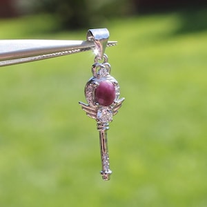May include: A silver pendant in the shape of a key, featuring a large, oval, deep red gemstone at the center. The pendant is adorned with small, clear gemstones and wing-like details, with a crown-like design at the top.