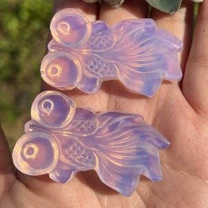 May include: Two opalescent crystal goldfish figurines. The fish have large, round eyes and flowing tail fins. The colors range from pale pink to lavender, with iridescent highlights. The fish are carved with detailed scales and fins.