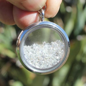 May include: A circular silver-toned metal locket pendant featuring a clear glass front and back. The interior of the locket is filled with numerous small, clear, faceted, and irregular-shaped crystals that move freely within the casing.