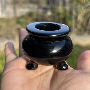May include: A small, glossy black cauldron-shaped object with three rounded feet and a circular opening at the top. The smooth, polished surface reflects light, highlighting its rounded form. This decorative piece is likely used for rituals or as a decorative item.