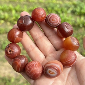 May include: A bracelet of polished, round agate beads in shades of red, brown, and orange. The beads have unique patterns of swirls and bands. The bracelet is held in a hand against a blurred green background.