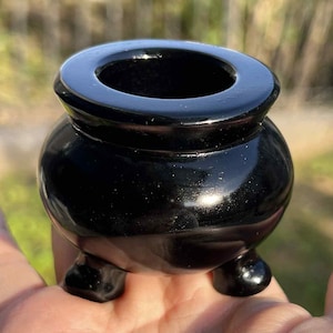 May include: A small, glossy black cauldron with three rounded feet. The cauldron has a wide, circular opening and a rounded body. The surface reflects light, and the cauldron appears to be made of a solid, dark material. The background is blurred.