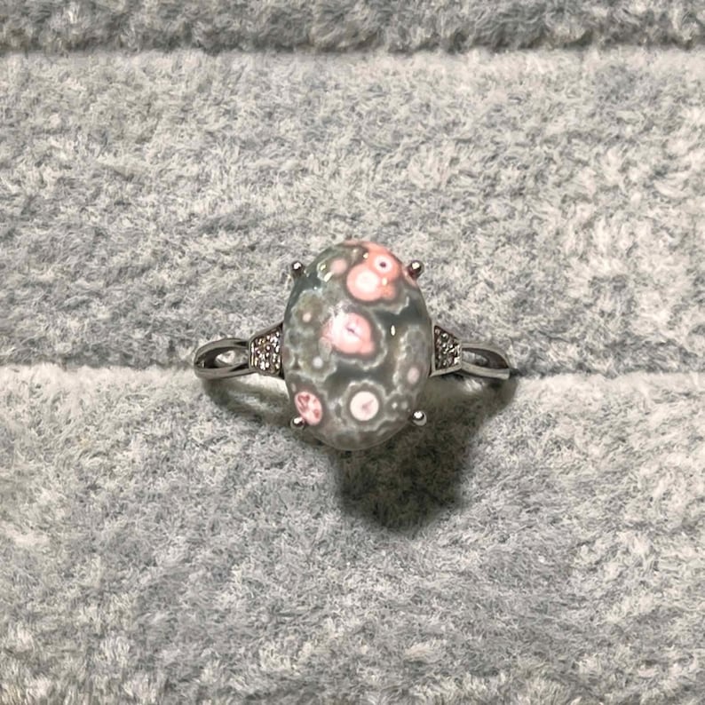 Peut inclure: Une bague argent&eacute;e avec une pierre cabochon ovale &agrave; base grise et motifs circulaires roses. L'anneau en m&eacute;tal comporte de petites pierres claires serties de chaque c&ocirc;t&eacute; de la monture centrale, maintenue par quatre griffes en m&eacute;tal.
