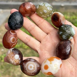 May include: A collection of polished agate stones in various colors, including brown, white, black, and green. The stones have unique patterns and textures, held in a hand. The stones are oval and round.