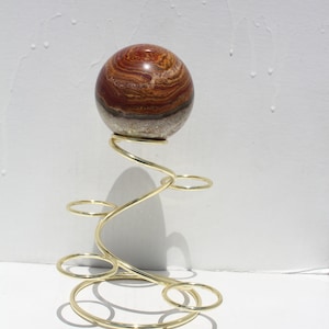 May include: A decorative sculpture featuring a polished, spherical stone with bands of red, brown, and tan hues. The stone rests atop a gold-colored, swirling metal stand with multiple circular accents, creating a modern, artistic display.