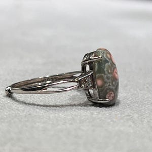 Peut inclure: Une bague argent&eacute;e orn&eacute;e d'une pierre ovale aux motifs tachet&eacute;s verts et roses, fix&eacute;e dans une monture en m&eacute;tal &agrave; griffes. L'anneau est divis&eacute; pr&egrave;s du sertissage et comporte une petite pierre transparente de chaque c&ocirc;t&eacute;, avec le poin&ccedil;on S925.