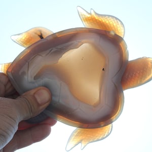 May include: A translucent, carved stone turtle figurine. The turtle has a brown and tan body with detailed flipper and head designs. The figurine is held against a bright, white background.