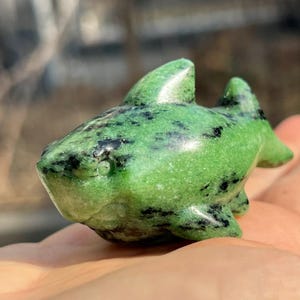 May include: A small, green stone shark figurine with black speckles. The shark has a detailed fin and tail design. The figurine is a decorative item, possibly a collectible or a unique piece of home decor.