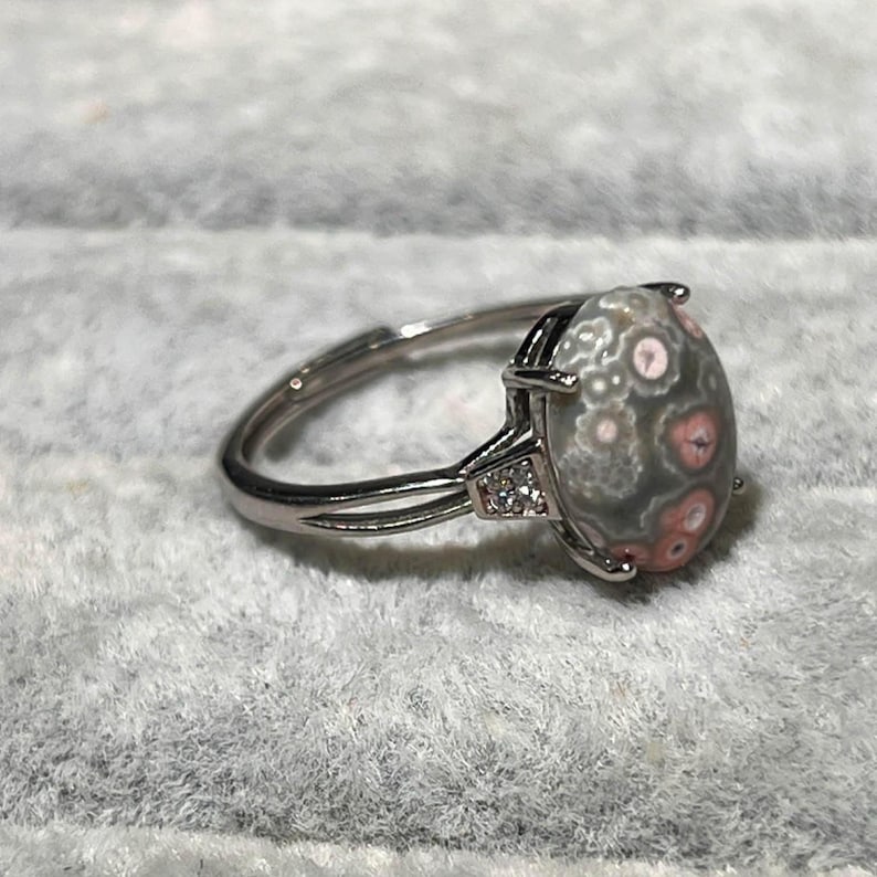 Peut inclure: Une bague argent&eacute;e orn&eacute;e d'une pierre cabochon ovale pr&eacute;sentant un motif orbiculaire gris et rose. La monture pr&eacute;sente un design &agrave; anneau divis&eacute; avec de petites pierres d'accent transparentes incrust&eacute;es de chaque c&ocirc;t&eacute; de la pierre centrale.