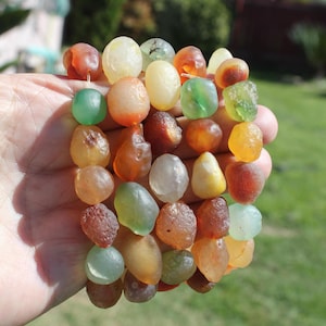 May include: A bracelet featuring a collection of colorful, irregularly shaped beads. The beads are in shades of green, yellow, orange, and brown. The bracelet is held in a hand against a blurred green background.