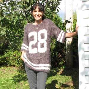 May include: A woman wearing a brown fuzzy sweater with white stripes and the number "28" on the front. She is wearing dark gray jeans.
