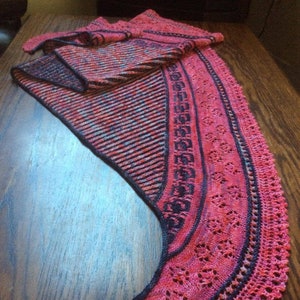 May include: A pink and black knitted shawl with a lacy border. The shawl is folded in half and laid on a wooden surface.