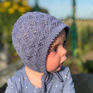 May include: A light blue knitted baby bonnet with a delicate openwork pattern. The bonnet has a rounded shape and ties under the chin with matching blue cords. The baby is wearing a blue onesie with white star patterns.