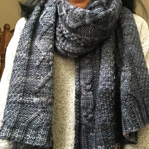 Puede incluir: Una bufanda de punto azul con un patr&oacute;n de trenza. La bufanda est&aacute; doblada por la mitad y drapeada alrededor del cuello de una persona.