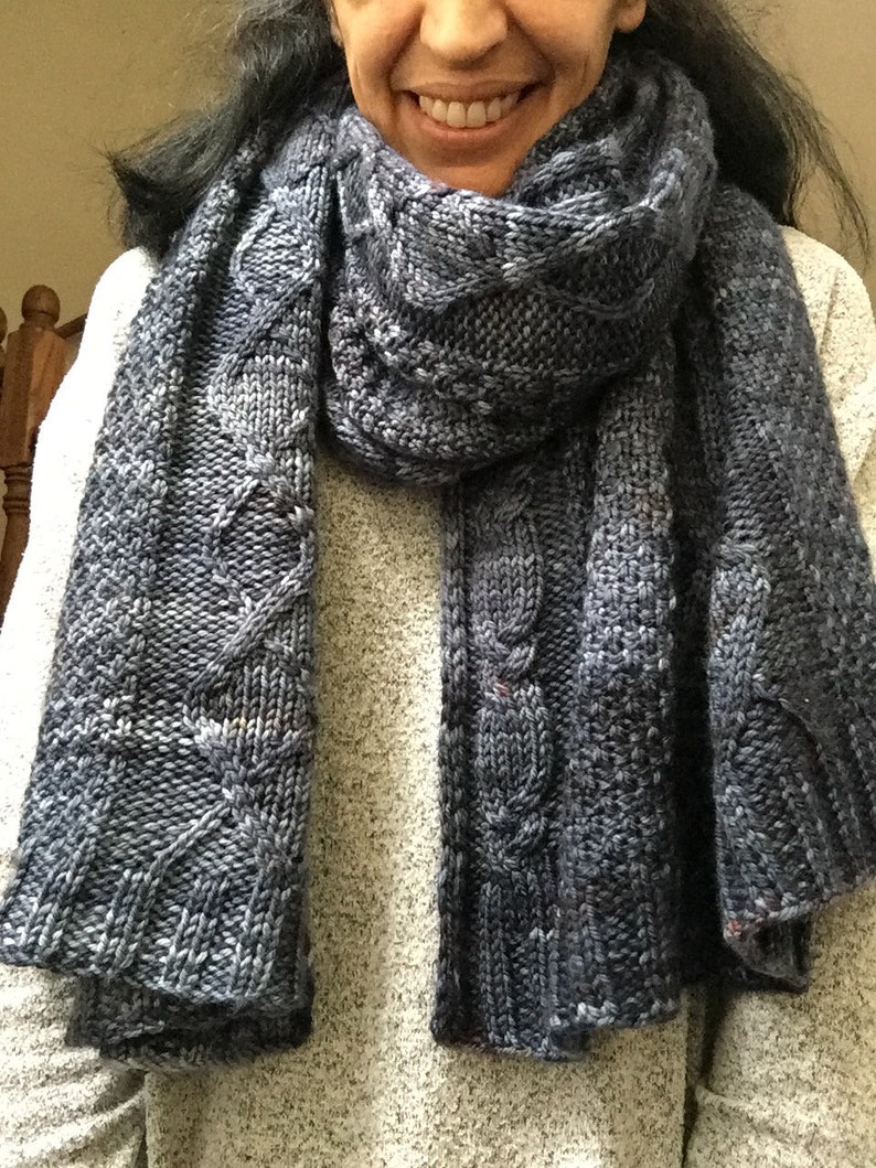 Puede incluir: Una bufanda de punto azul con un patr&oacute;n de trenza. La bufanda est&aacute; doblada por la mitad y drapeada alrededor del cuello de una persona.