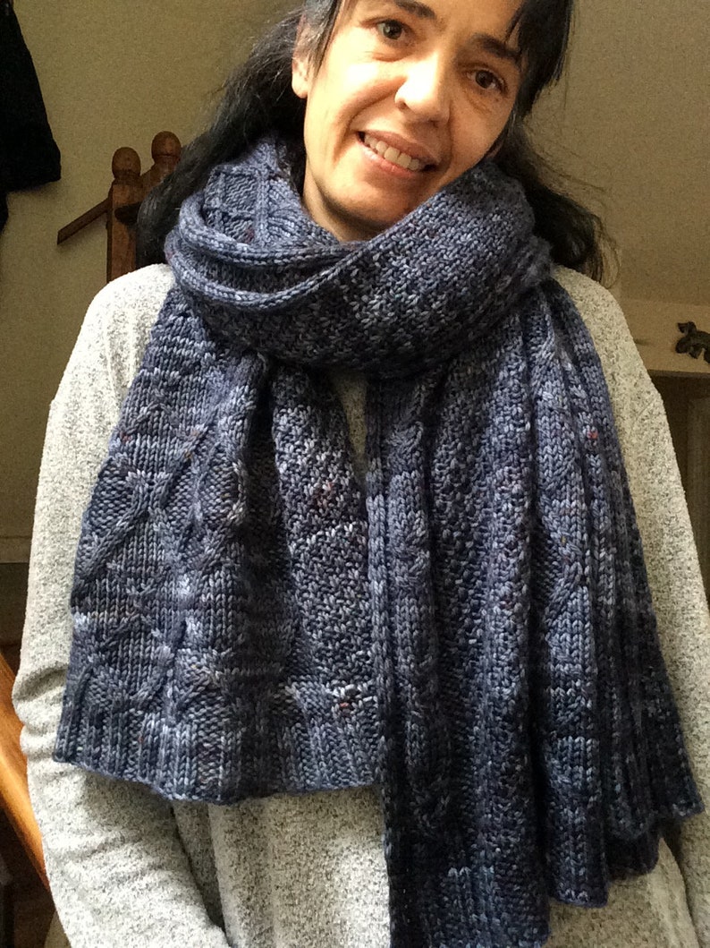 Puede incluir: Una bufanda de punto azul con un patr&oacute;n de trenza. La bufanda la lleva puesta una persona con el pelo oscuro.