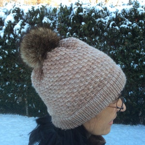 Peut inclure: Un bonnet en tricot avec un pompon en fourrure marron. Le bonnet est de couleur marron clair et présente un motif texturé.