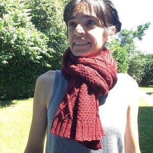 Puede incluir: Una mujer con una camisa gris sin mangas y una bufanda de punto roja alrededor del cuello. La bufanda es de punto de cable.