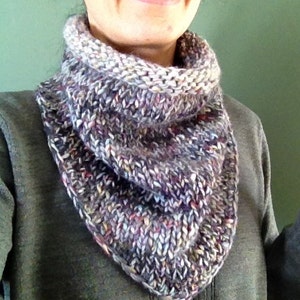 May include: A close-up of a woman wearing a gray knitted cowl neck scarf with a multi-colored pattern. The scarf is made of a soft, textured yarn and has a loose, drapey fit.