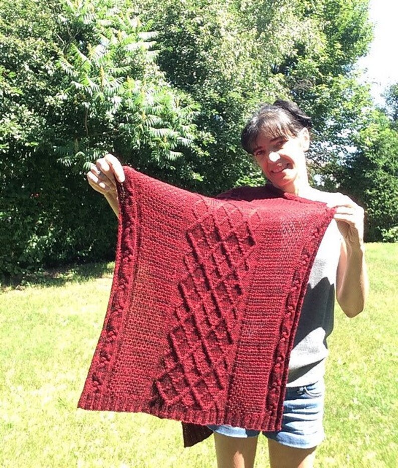 Puede incluir: Un chal de punto rojo con un patr&oacute;n geom&eacute;trico. El chal est&aacute; siendo sostenido por una persona frente a un c&eacute;sped verde.