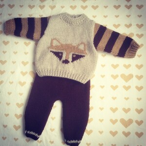 May include: A knitted baby outfit with a brown and beige striped sweater featuring a raccoon face and brown pants.