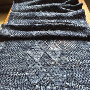 Puede incluir: Una bufanda de punto azul con un patrón de diamantes. La bufanda está doblada por la mitad y tiene una superficie texturizada.