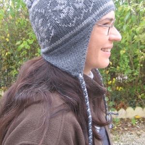 May include: A gray knit hat with ear flaps and a pom-pom. The hat features a white snowflake design and braided ties. The person in the image is wearing glasses and a brown jacket.