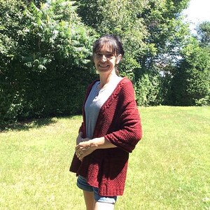 Puede incluir: Una mujer lleva un c&aacute;rdigan de punto rojo con una camisa gris debajo y pantalones cortos de mezclilla. Est&aacute; de pie en un &aacute;rea cubierta de hierba con &aacute;rboles en el fondo.