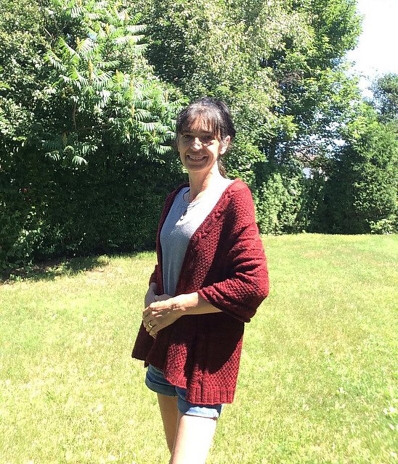 Puede incluir: Una mujer lleva un c&aacute;rdigan de punto rojo con una camisa gris debajo y pantalones cortos de mezclilla. Est&aacute; de pie en un &aacute;rea cubierta de hierba con &aacute;rboles en el fondo.