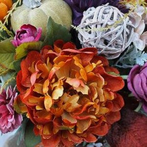May include: A floral arrangement featuring a large burnt orange peony, deep purple roses, and smaller orange blossoms. A light green decorative pumpkin and a white woven twig sphere are nestled among the flowers, accented by green leaves.