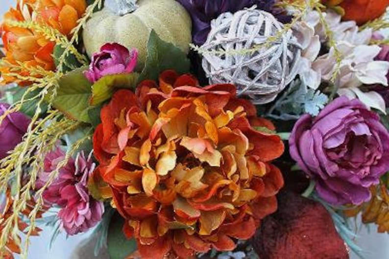 May include: A floral arrangement featuring a large burnt orange peony, deep purple roses, and smaller orange blossoms. A light green decorative pumpkin and a white woven twig sphere are nestled among the flowers, accented by green leaves.