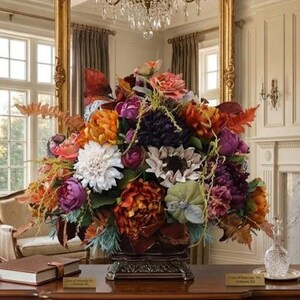 May include: A large, dense floral arrangement featuring a mix of artificial flowers in shades of deep purple, burnt orange, white, and muted green. The bouquet includes peonies, dahlias, and ferns, all contained within a dark, ornate pedestal base.
