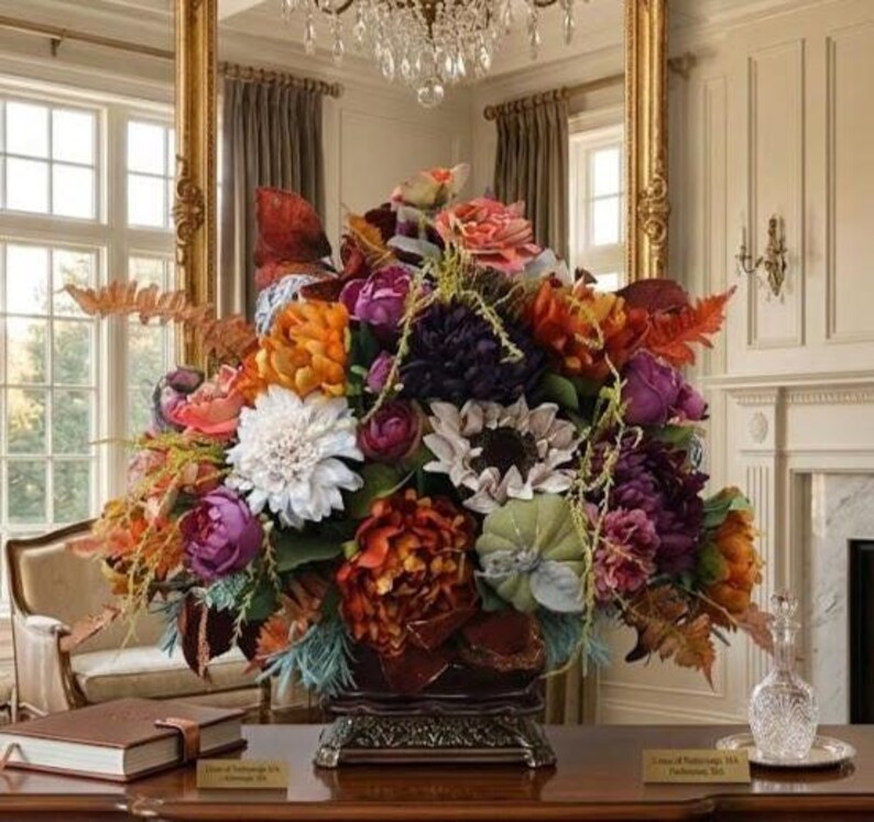 May include: A large, dense floral arrangement featuring a mix of artificial flowers in shades of deep purple, burnt orange, white, and muted green. The bouquet includes peonies, dahlias, and ferns, all contained within a dark, ornate pedestal base.